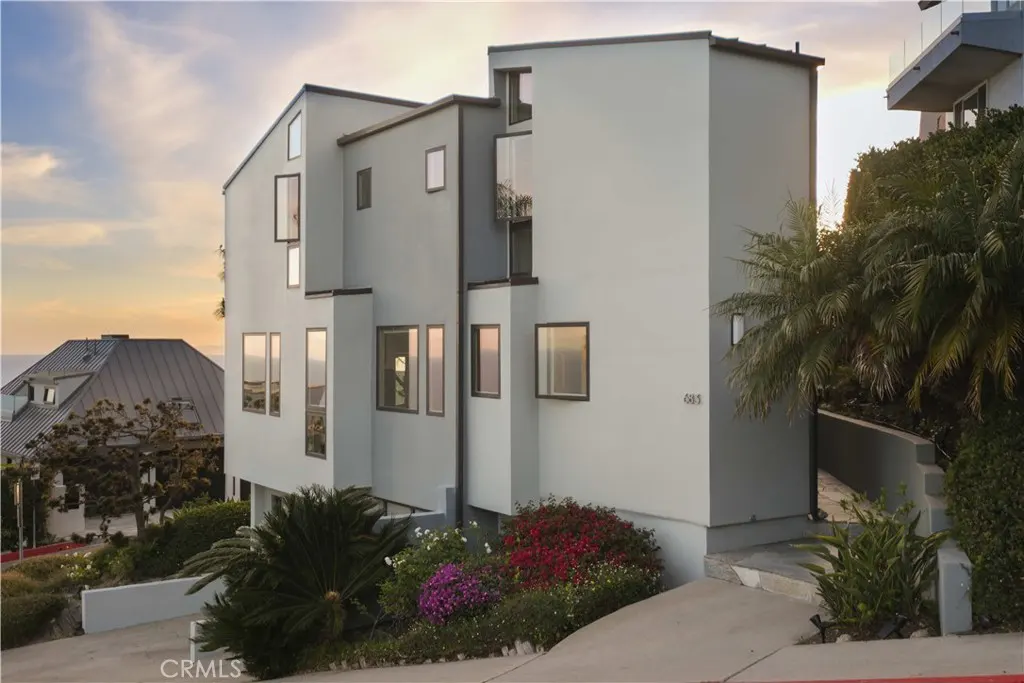 Modern three-story gray house with many windows, landscaping, and a driveway. Ocean and sunset in the background.