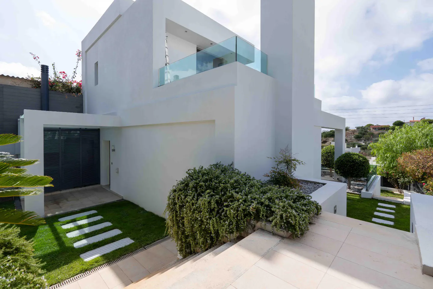 Modern white house exterior with a gray garage door, green lawn, and stone walkway. A glass balcony is visible on the second floor.