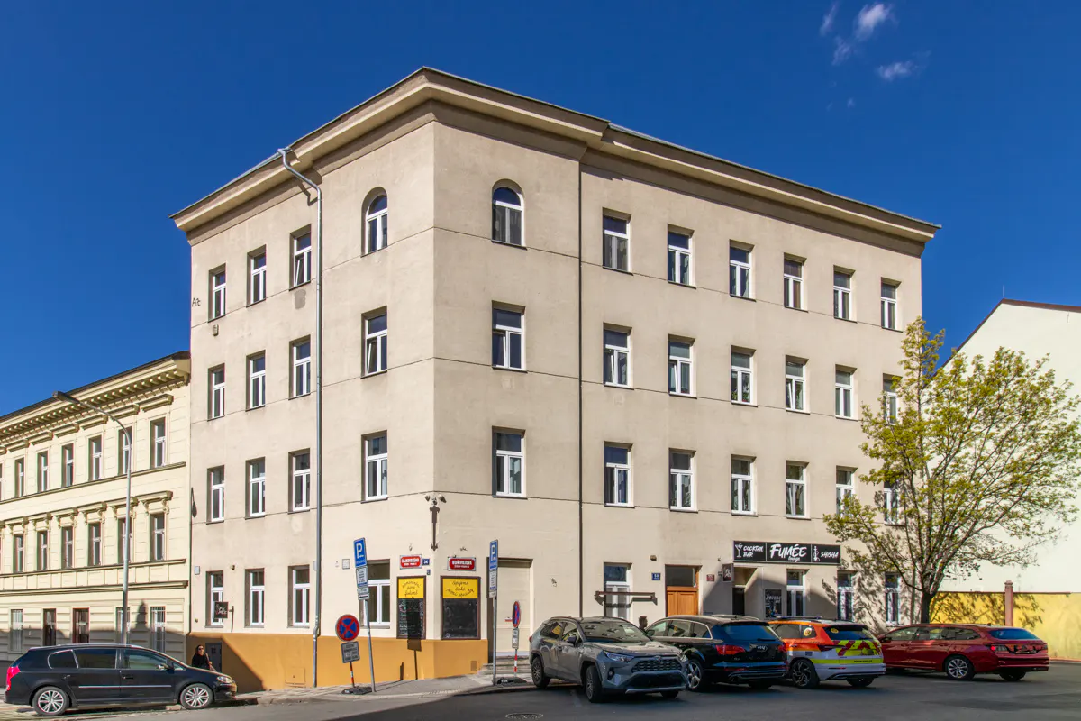 A four-story beige building with white-framed windows under a blue sky, cars parked in front.