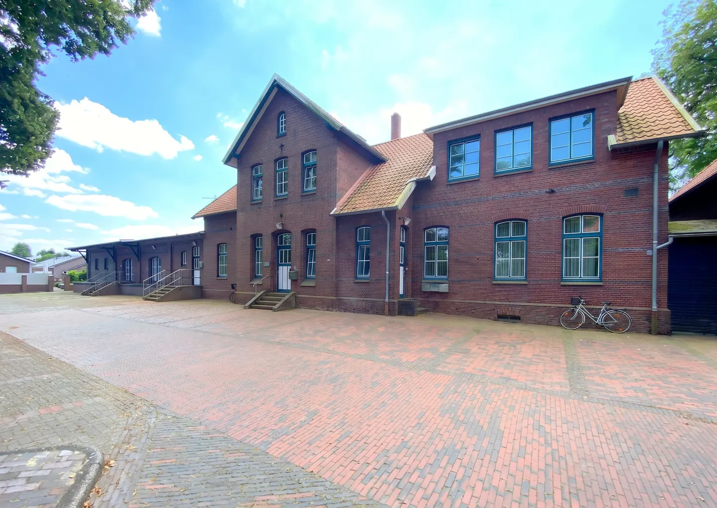 A two-story red brick building with a red tile roof and blue window frames. A bicycle is parked against the building.