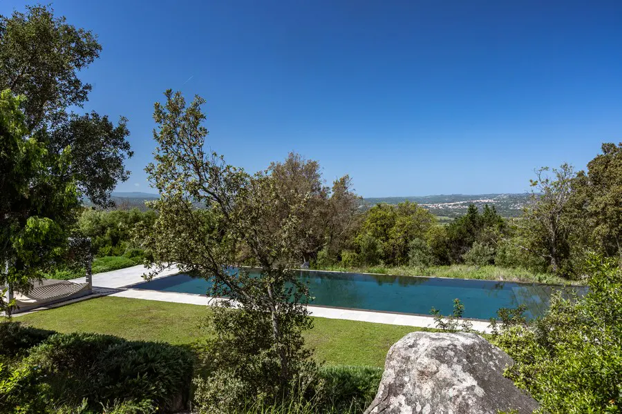 Scenic view of a long, rectangular pool surrounded by green grass and trees under a clear blue sky. A town is visible in the distance.