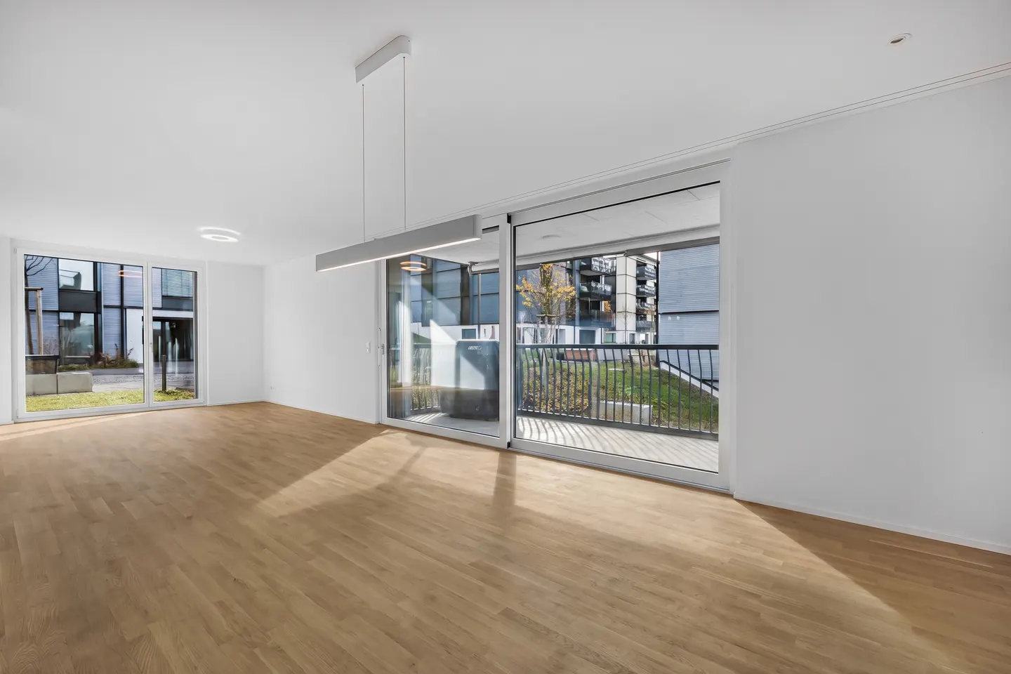 Bright, empty room with light wood floors, white walls, and large sliding glass doors to a balcony and courtyard.