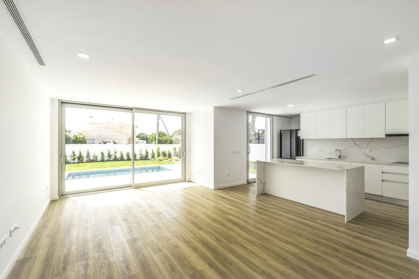 Bright, modern open-plan living space with wood floors, white walls, and a kitchen island. Sliding glass doors lead to a pool.