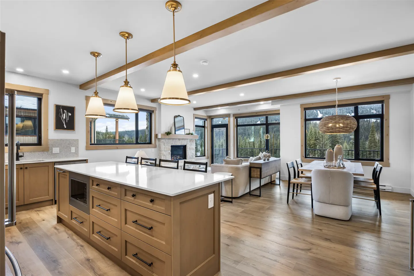 Open-concept living space with a kitchen island, dining table, and living room. Large windows offer views of a forested mountain landscape.
