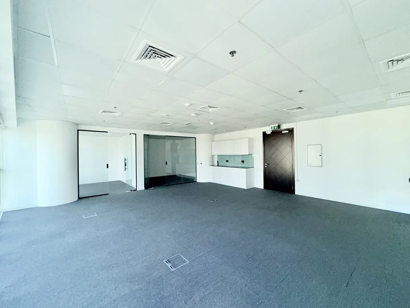 Empty office space with gray carpet, white walls, and a white ceiling with vents. There is a glass-walled room and a small kitchenette.