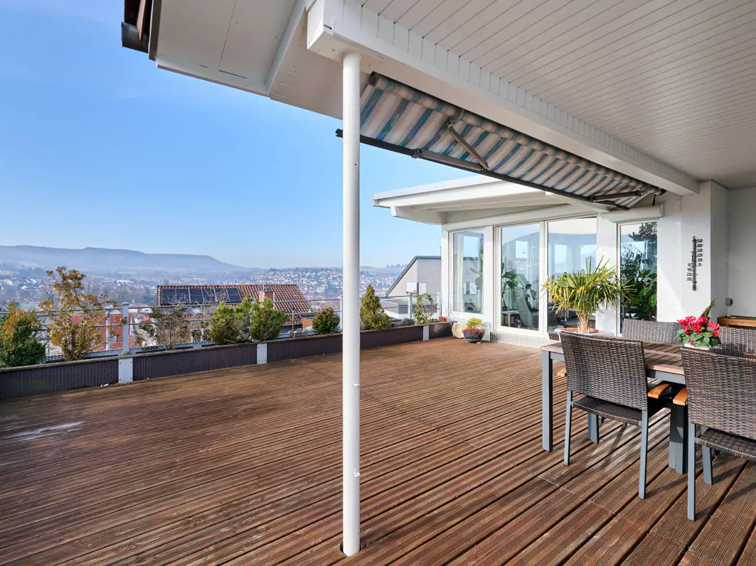 Wide wooden deck with table and chairs under a striped awning, overlooking a distant cityscape under a blue sky.