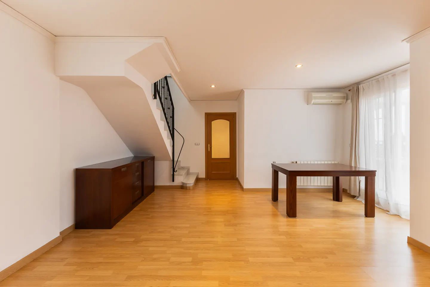 Bright, empty room with wood floors, white walls, and a staircase. A dark wood table and cabinet add contrast. Natural light streams through sheer curtains.