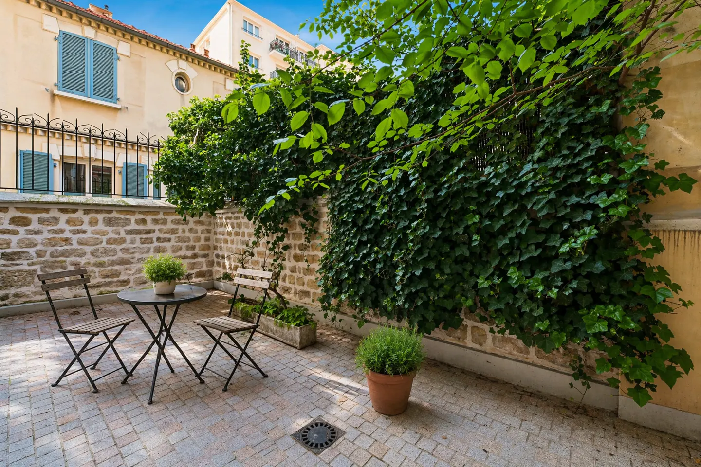 A courtyard with a stone wall covered in ivy, a round table, and two chairs. A potted plant sits on the stone-paved ground.