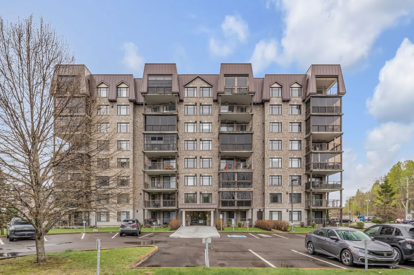 Exterior view of a tall, tan brick apartment building with balconies and a brown roof, parking lot in front.