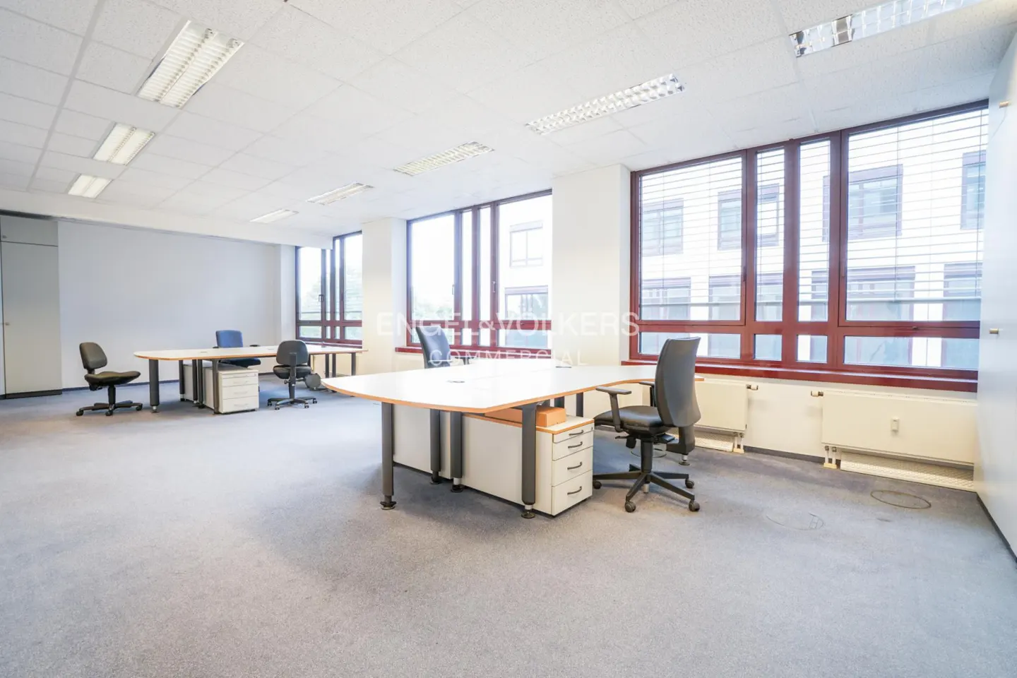 Bright, empty office space with desks, chairs, and large windows with brown frames. Gray carpet and white walls.