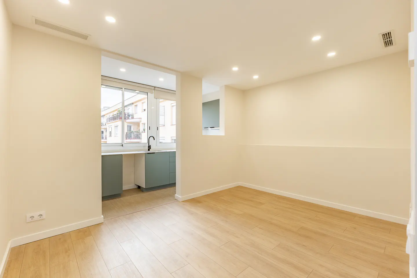 Bright, empty room with light wood floors and cream walls. A kitchen area with green cabinets is visible through a large opening.