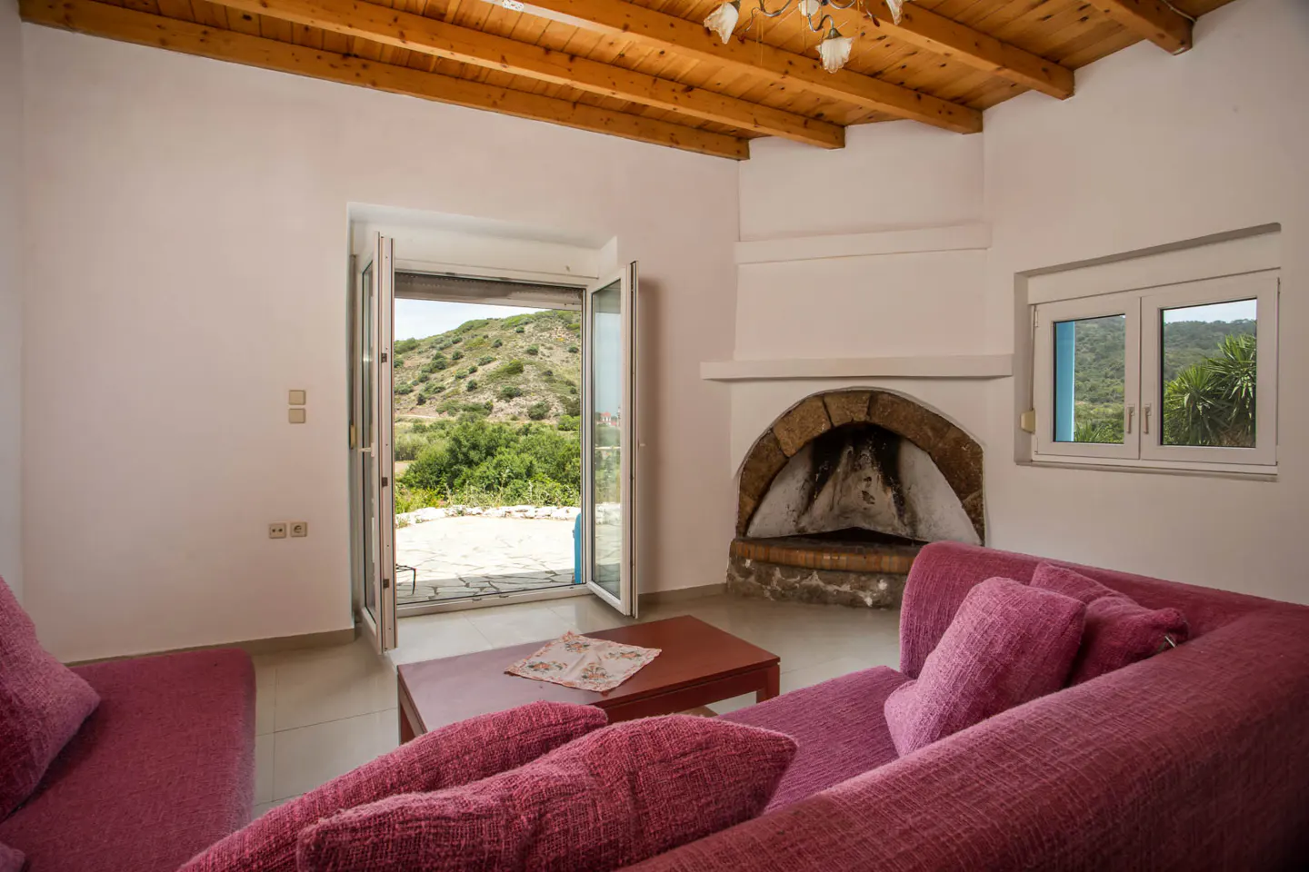 Living room with pink sofas, a stone fireplace, and an open door to a green hillside. The ceiling is wood.