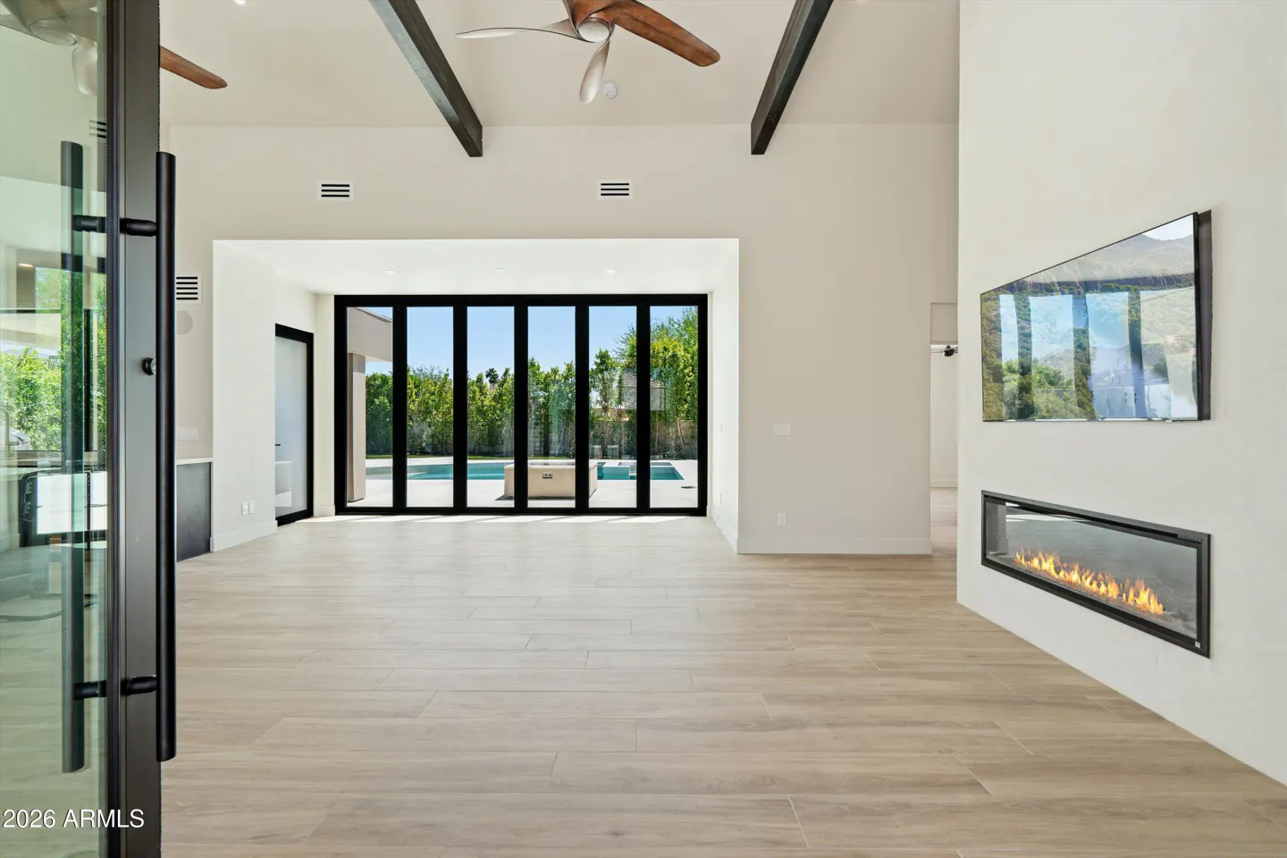 Bright, modern living room with wood floors, white walls, and black-framed folding doors leading to a pool. A linear fireplace and TV are on the right wall.