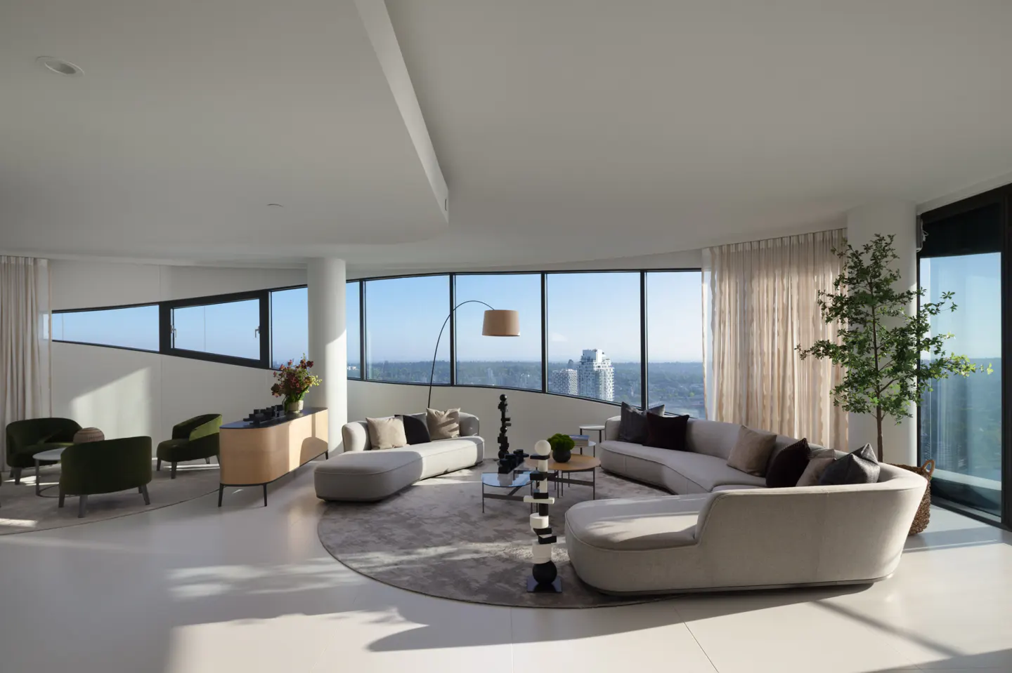 Bright, modern living room with curved sofas, floor lamp, and city views through large windows. A tree stands near the window.