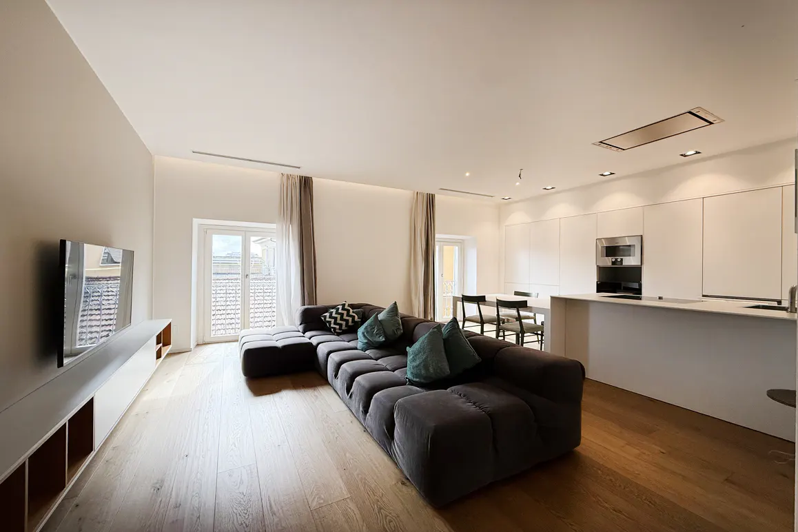 A modern, open-concept living space with a large, gray sectional sofa, a flat-screen TV, and a white kitchen with a breakfast bar.
