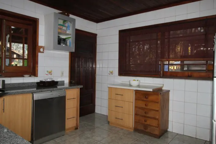 A kitchen with white tiled walls, wooden cabinets, and a dark wood ceiling. A window with brown blinds is visible.