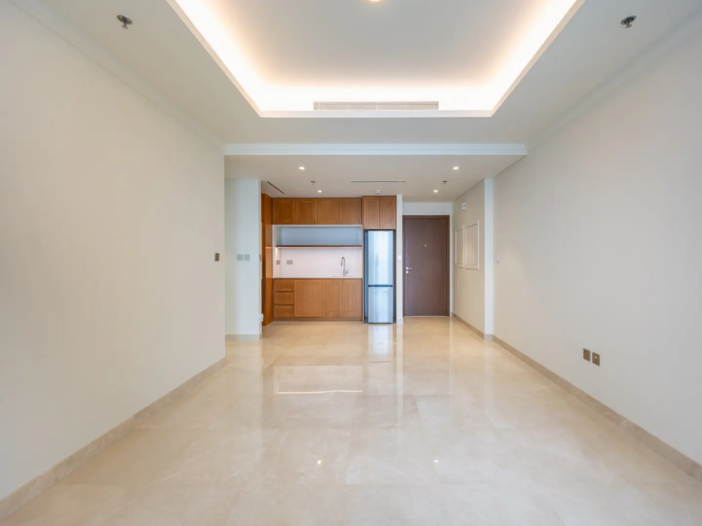 An empty apartment with white walls, a marble floor, and a small kitchen with wood cabinets and a stainless steel refrigerator.