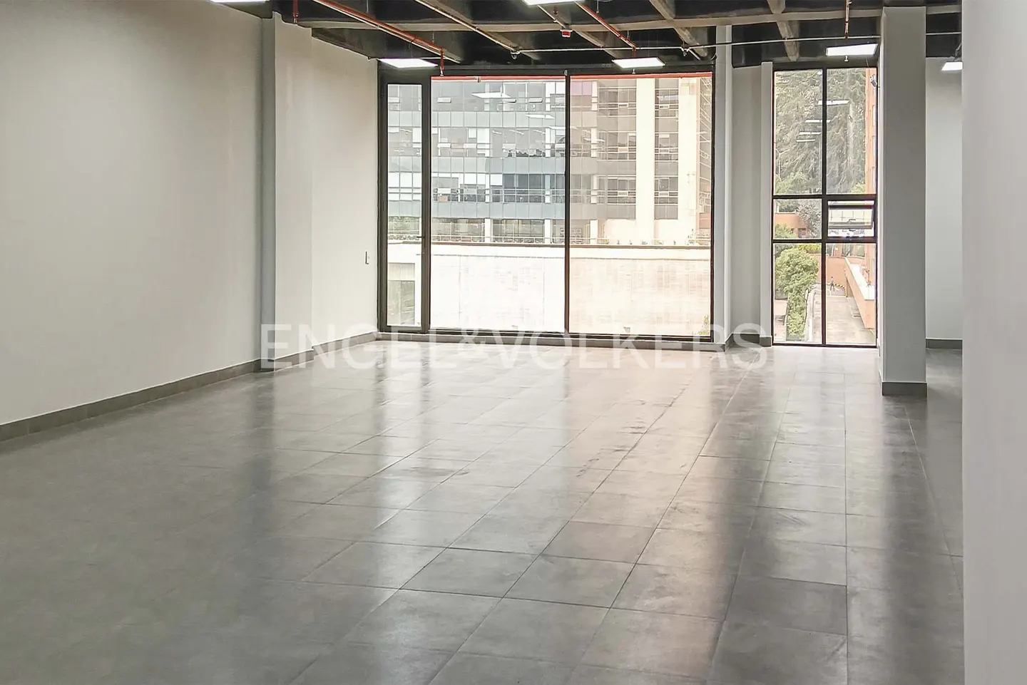 Empty office space with gray tiled floor, white walls, and large windows showing city buildings.