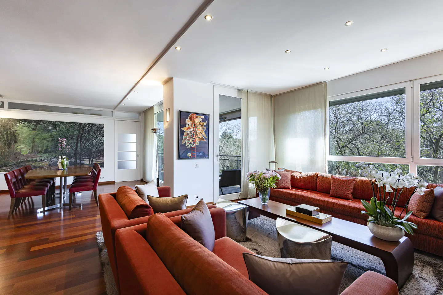 Bright living room with orange sofas, wood floors, and a dining table with red chairs. Large windows overlook trees.