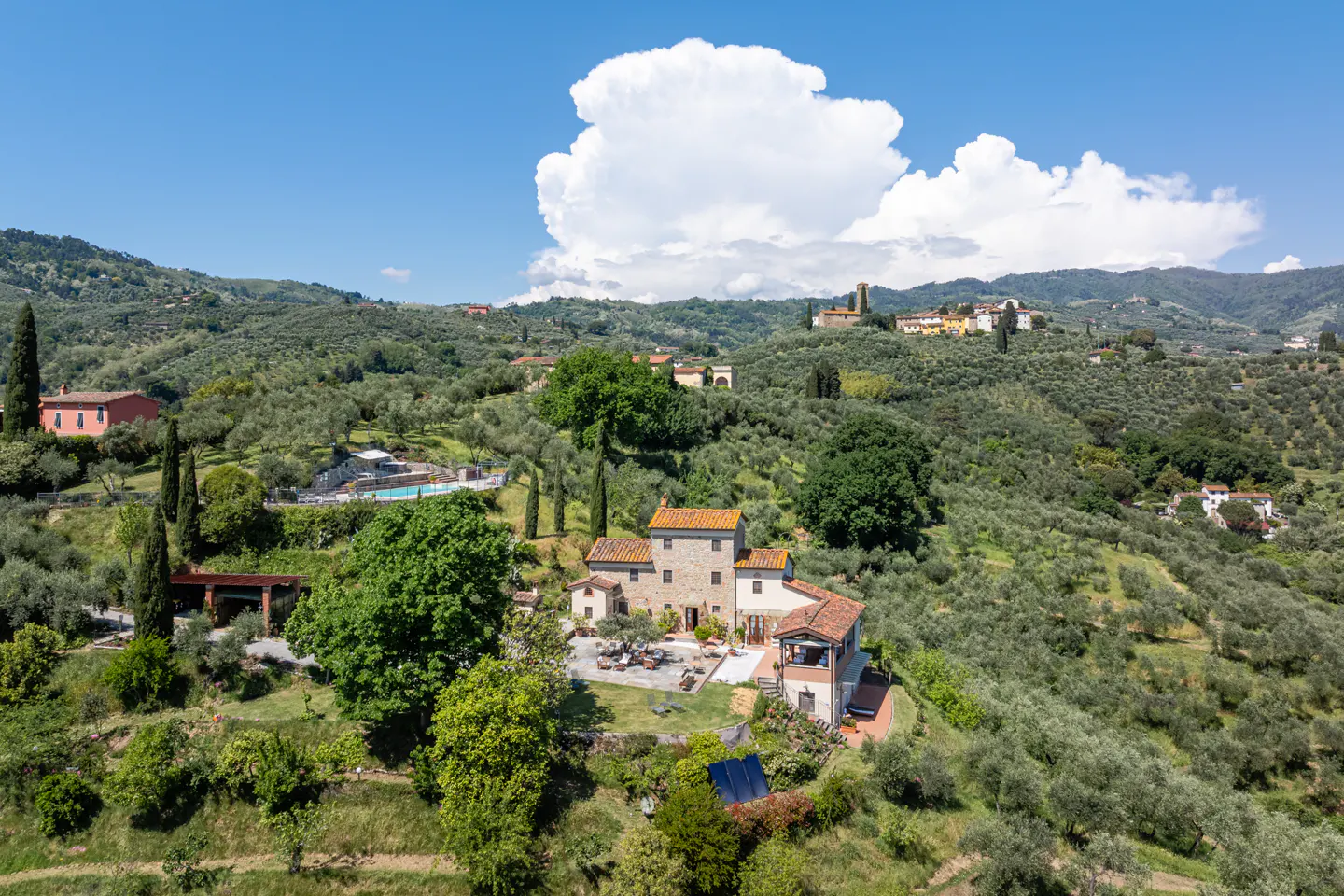 Aerial view of a Tuscan-style villa with a red tile roof, surrounded by olive trees and rolling hills under a blue sky with white clouds.