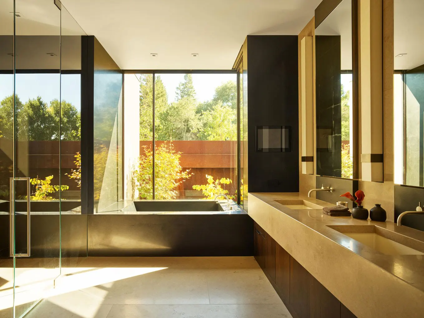 Modern bathroom with a large window overlooking greenery. Double sinks, dark cabinets, and a black accent wall with a fireplace.