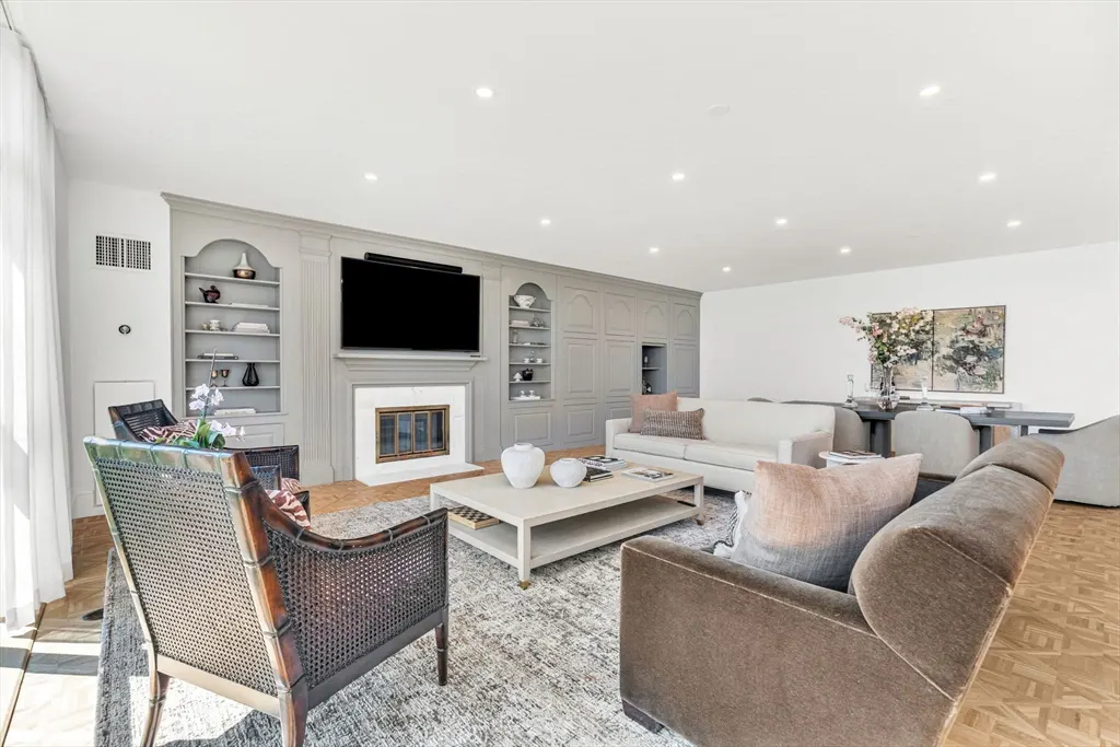 Bright living room with gray built-in shelves, fireplace, TV, sofas, and chairs on a patterned rug. A dining table is visible in the background.