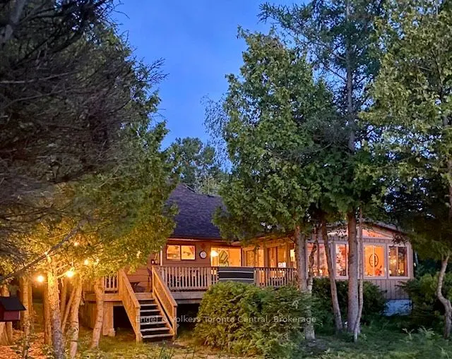 A cozy, wood-sided house with a deck is surrounded by trees at dusk, illuminated by warm interior lights and string lights.