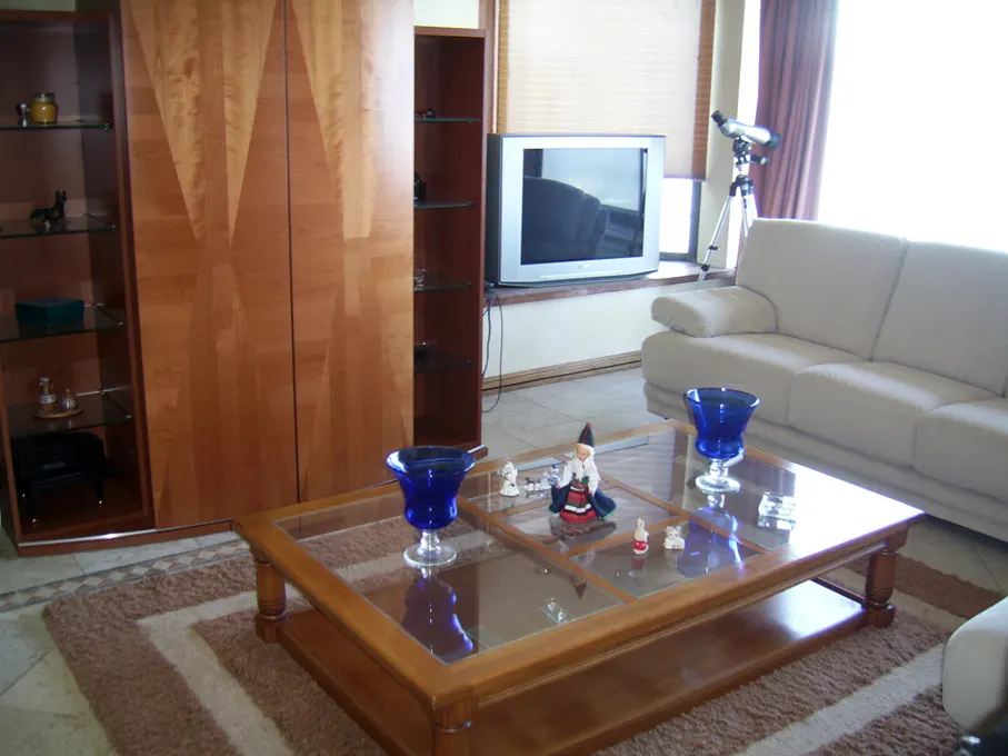 Living room with a glass-top coffee table, blue vases, and figurines. A white sofa, TV, and wooden cabinets are in the background.