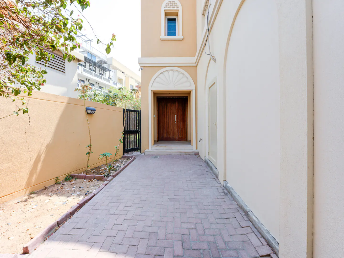 Exterior view of a tan house with a brick walkway leading to a wooden front door. A black gate is visible on the left.
