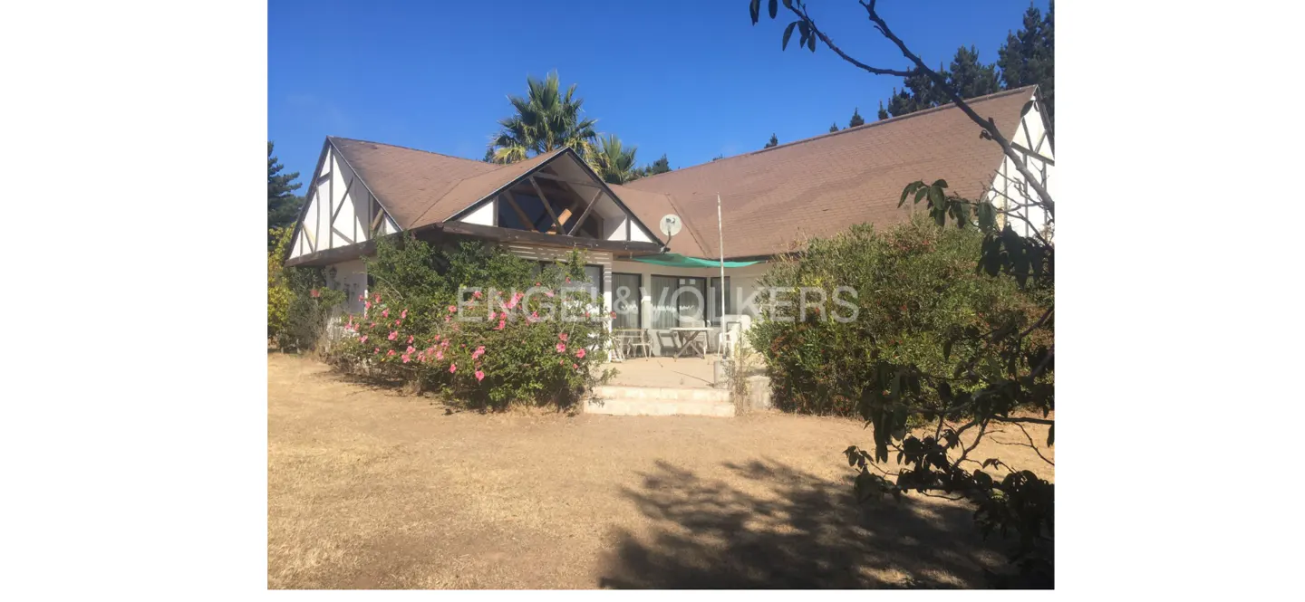 Exterior view of a white house with a brown roof, surrounded by greenery and a lawn. The Engel & Völkers logo is superimposed on the image.