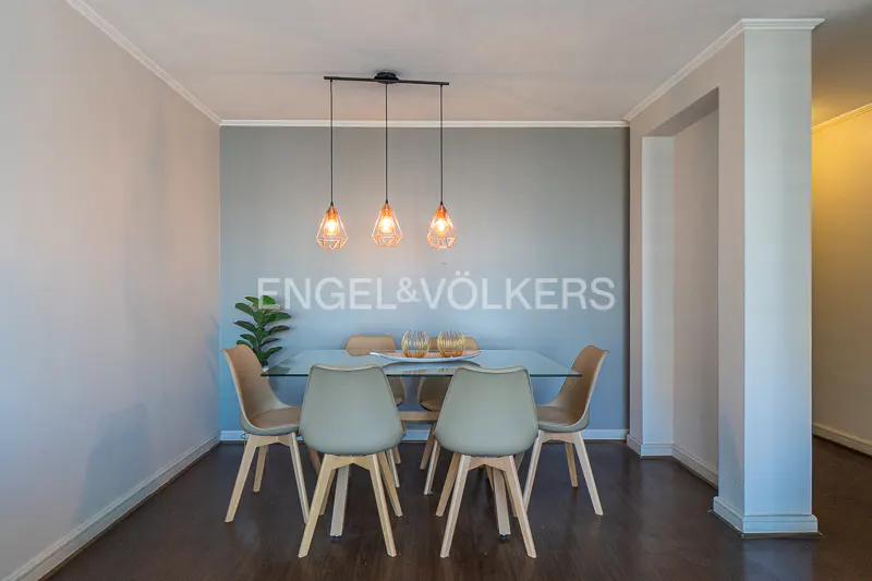 Dining room with glass table, six chairs, and three pendant lights. Engel & Völkers logo on the gray wall. Dark wood floor.