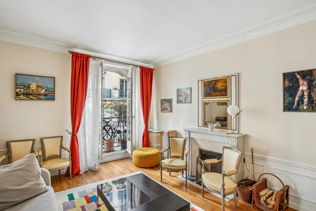 A bright living room with hardwood floors, cream walls, and red curtains framing an open balcony. Art adorns the walls above a fireplace and chairs.