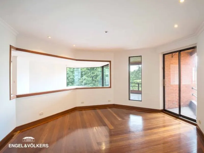 Bright, empty room with hardwood floors, white walls, and large windows showing green trees. Sliding glass doors lead to a brick balcony.