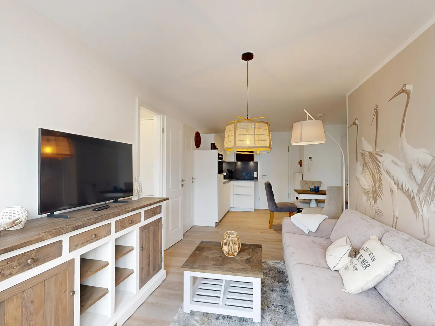 Living room with a TV on a wooden cabinet, a sofa with pillows, and a coffee table on a rug. A kitchen is visible in the background.