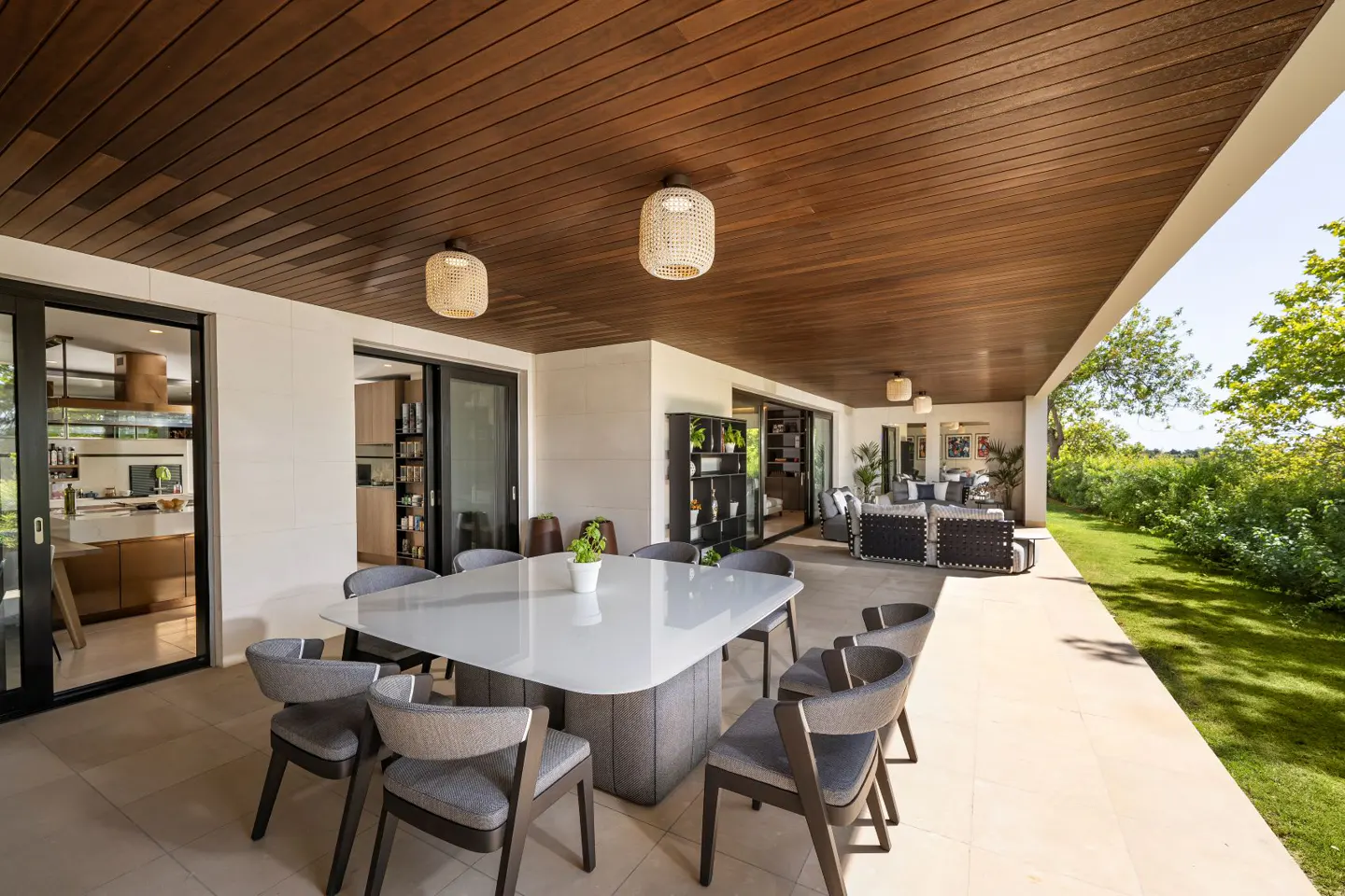 Outdoor dining area with a white table, gray chairs, and a wood ceiling with woven light fixtures. Sliding glass doors lead to the interior.