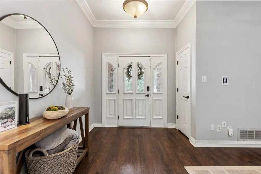 Foyer with dark wood floors, light gray walls, and a white front door with glass panels. A wooden console table with a round mirror sits to the left.