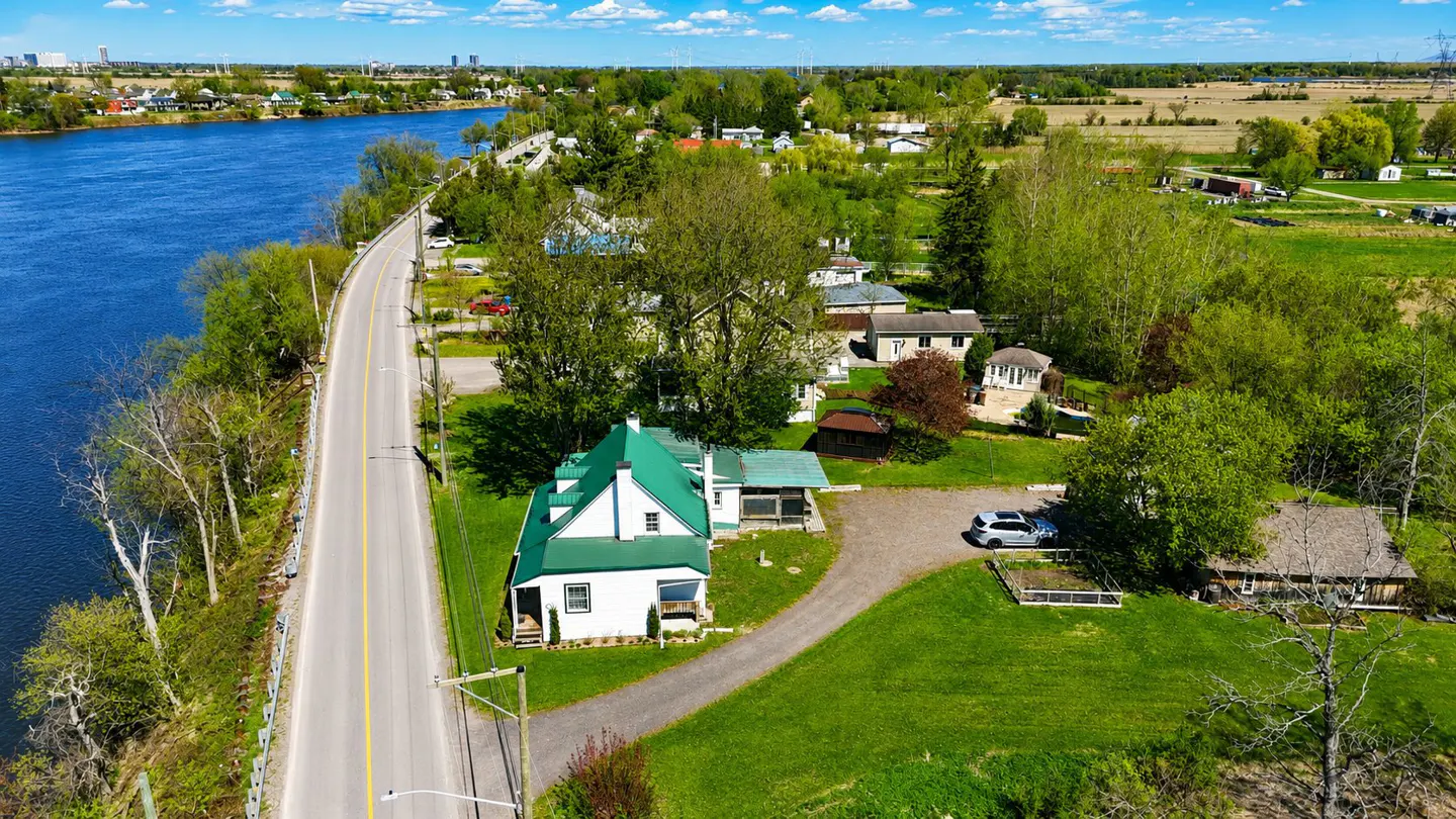 Aerial view of a white house with a green roof next to a road and a river.