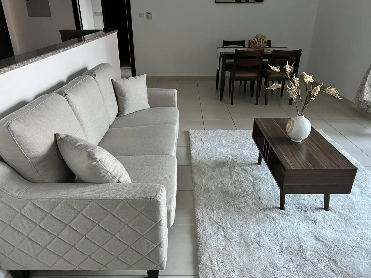 Living room with a beige sofa, a dark wood coffee table with a white vase, and a white fluffy rug. A dining table is in the background.