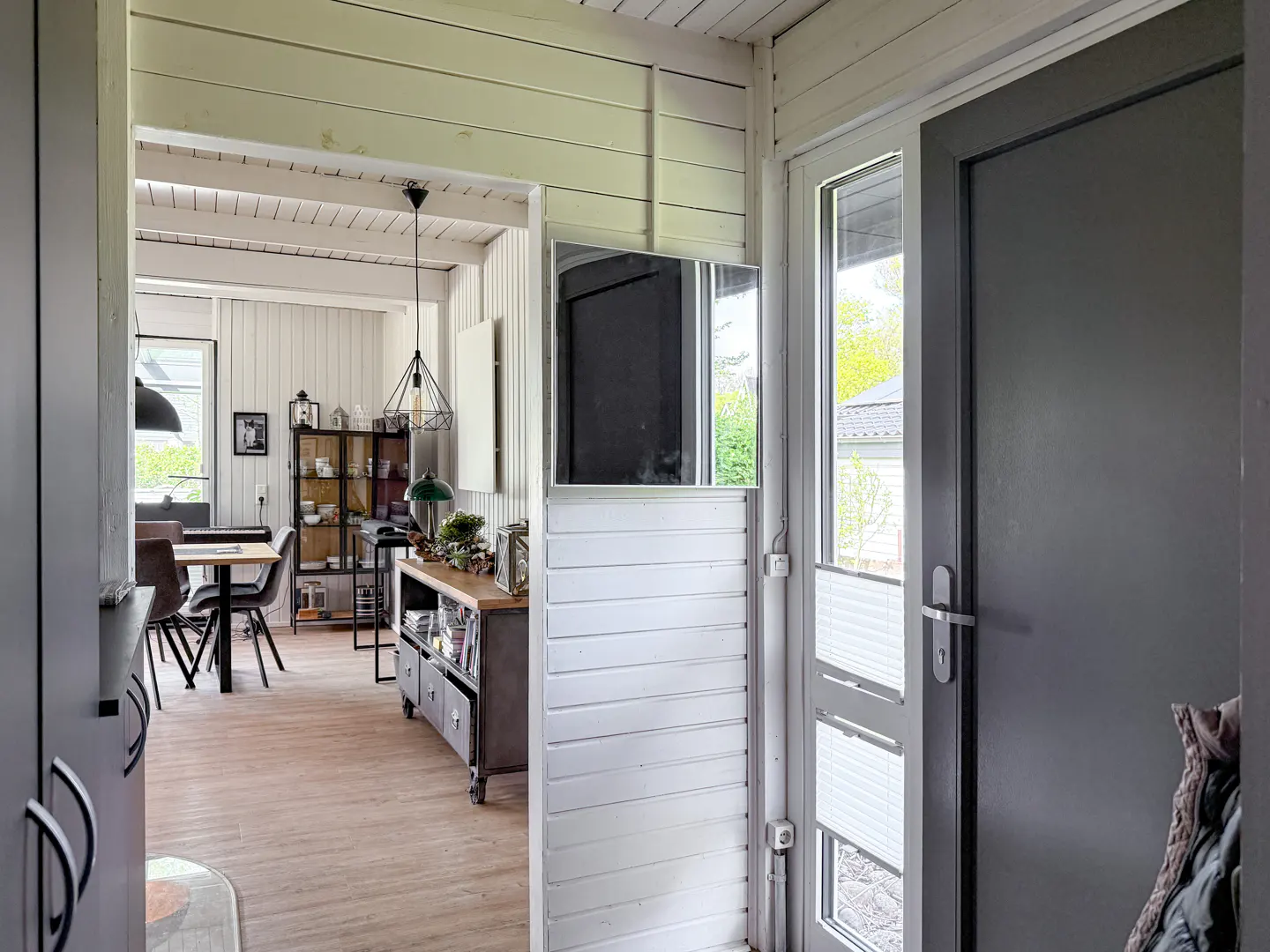 View from a grey entryway into a bright living area with white wood-paneled walls, a dining table, and industrial decor.