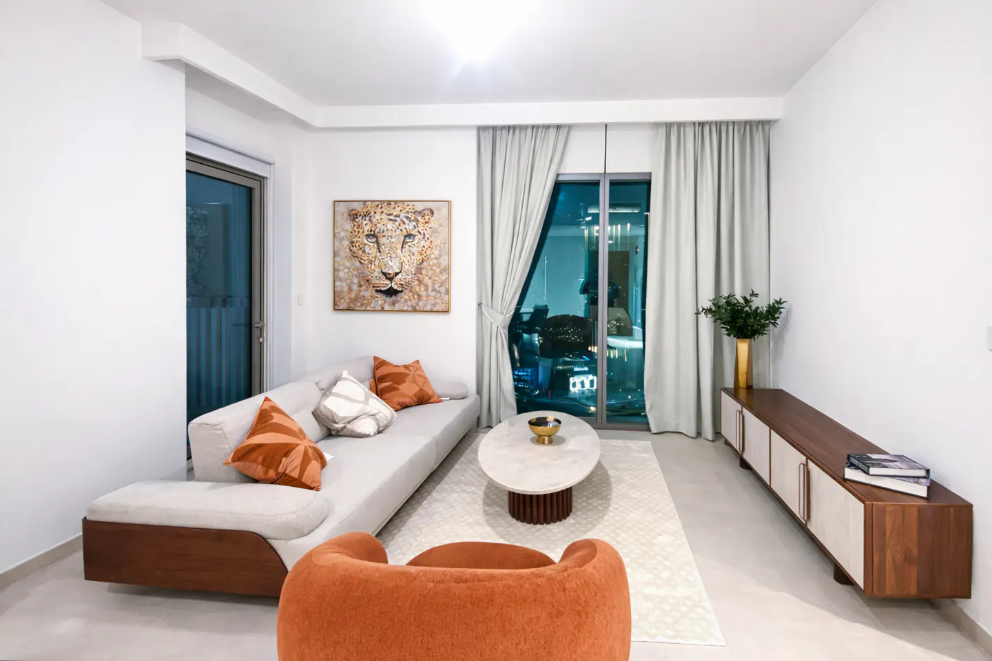 Bright living room with white walls, a gray sofa with orange pillows, and a leopard art piece. A round coffee table sits on a white rug.
