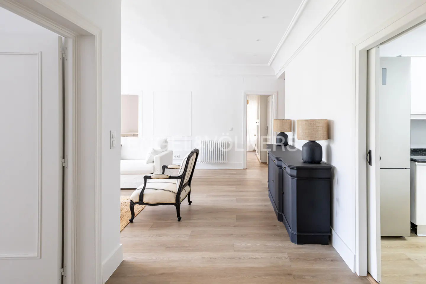 Bright, airy hallway with light wood floors, white walls, and a black sideboard with lamps. A striped chair sits near a doorway to a living room.