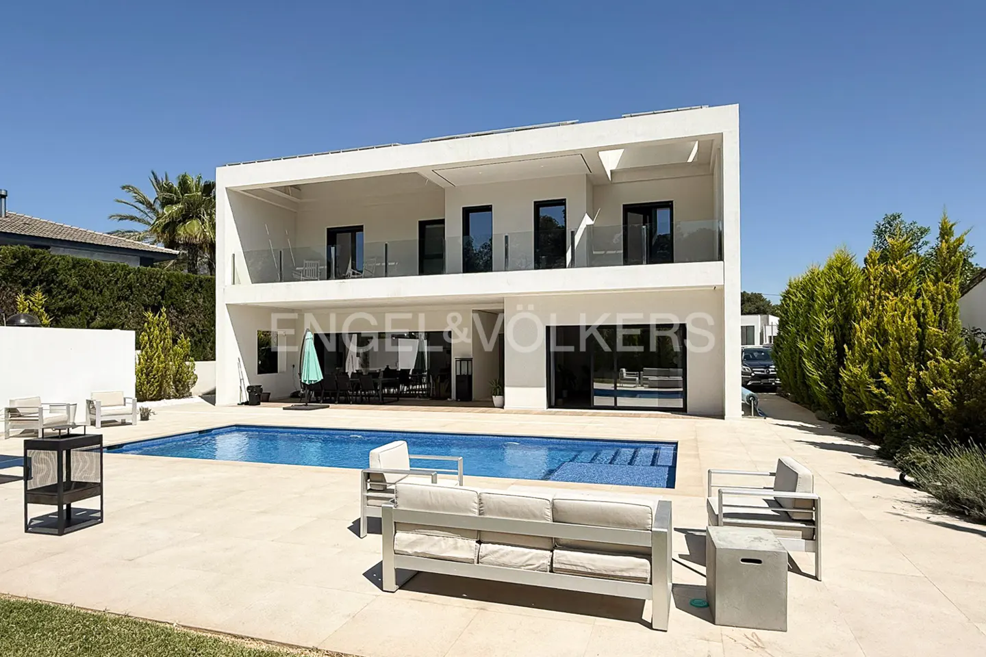Two-story white modern house with a blue pool and outdoor seating on a sunny day.