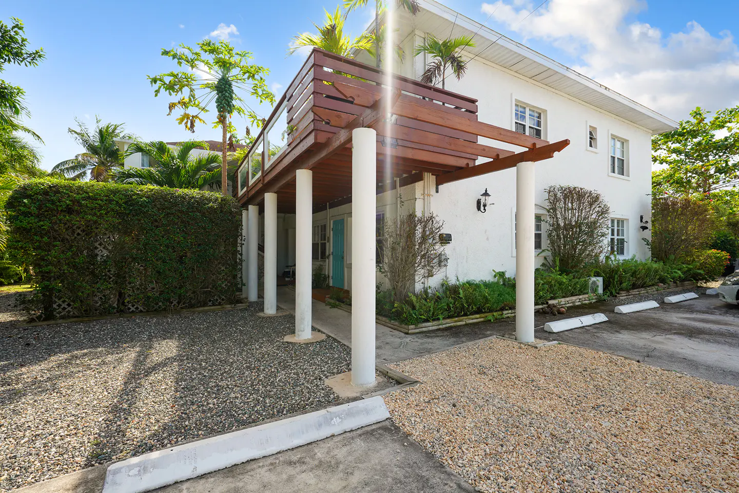 Exterior of a white two-story building with a wood balcony supported by white columns. Tropical plants and gravel surround the building.