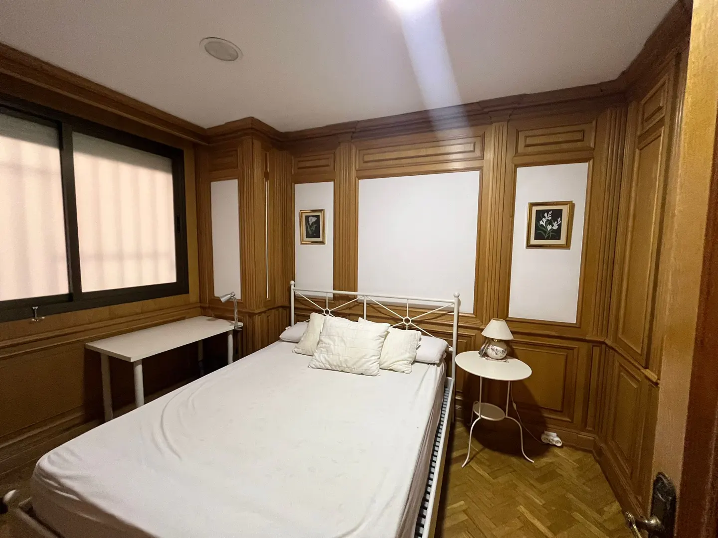 Bedroom with white bed, wood-paneled walls, desk, and side table on a herringbone wood floor.