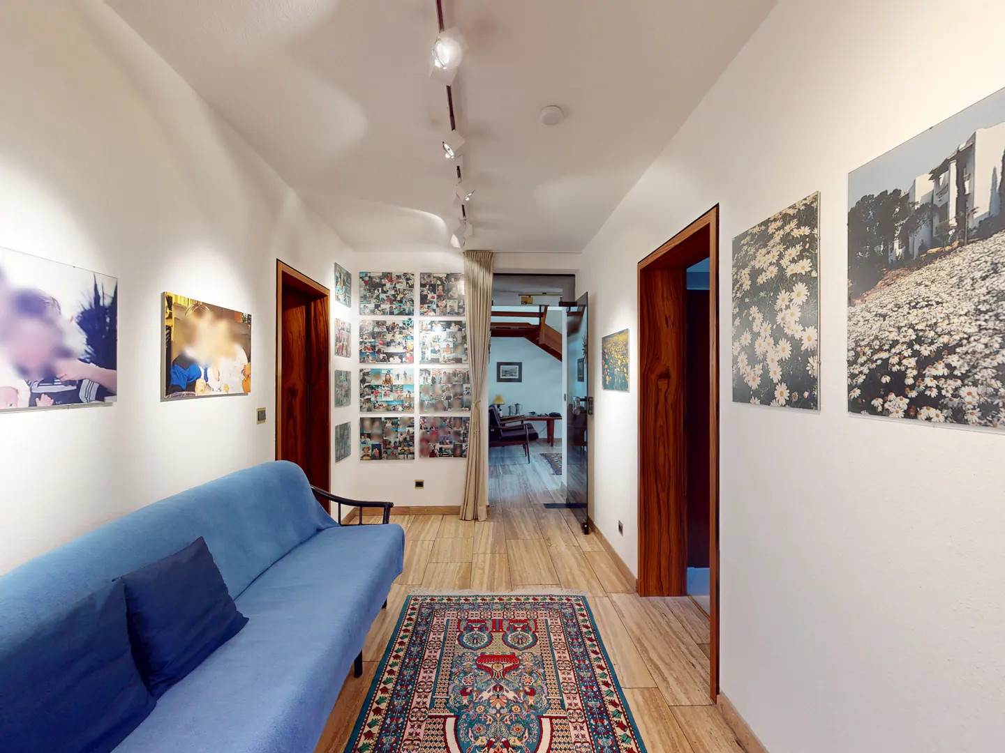 Hallway with white walls, wood floors, and art. A blue sofa sits on the left, and a colorful rug is on the floor.