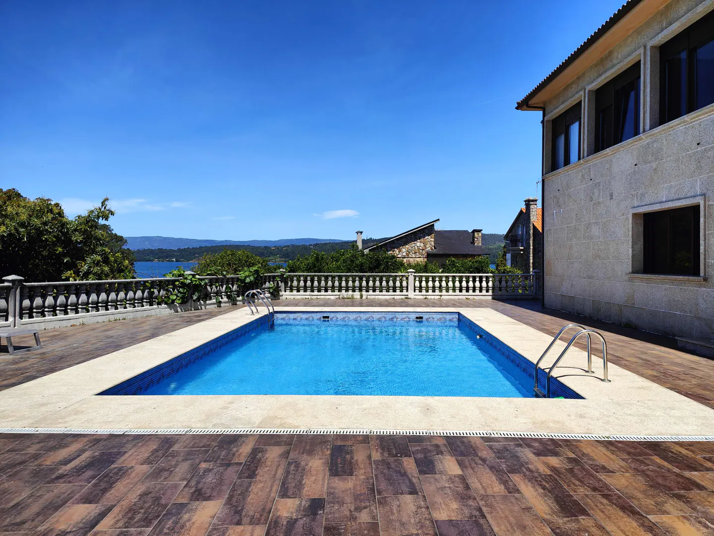 A rectangular swimming pool with blue water sits on a tiled patio next to a stone house, overlooking a distant bay.
