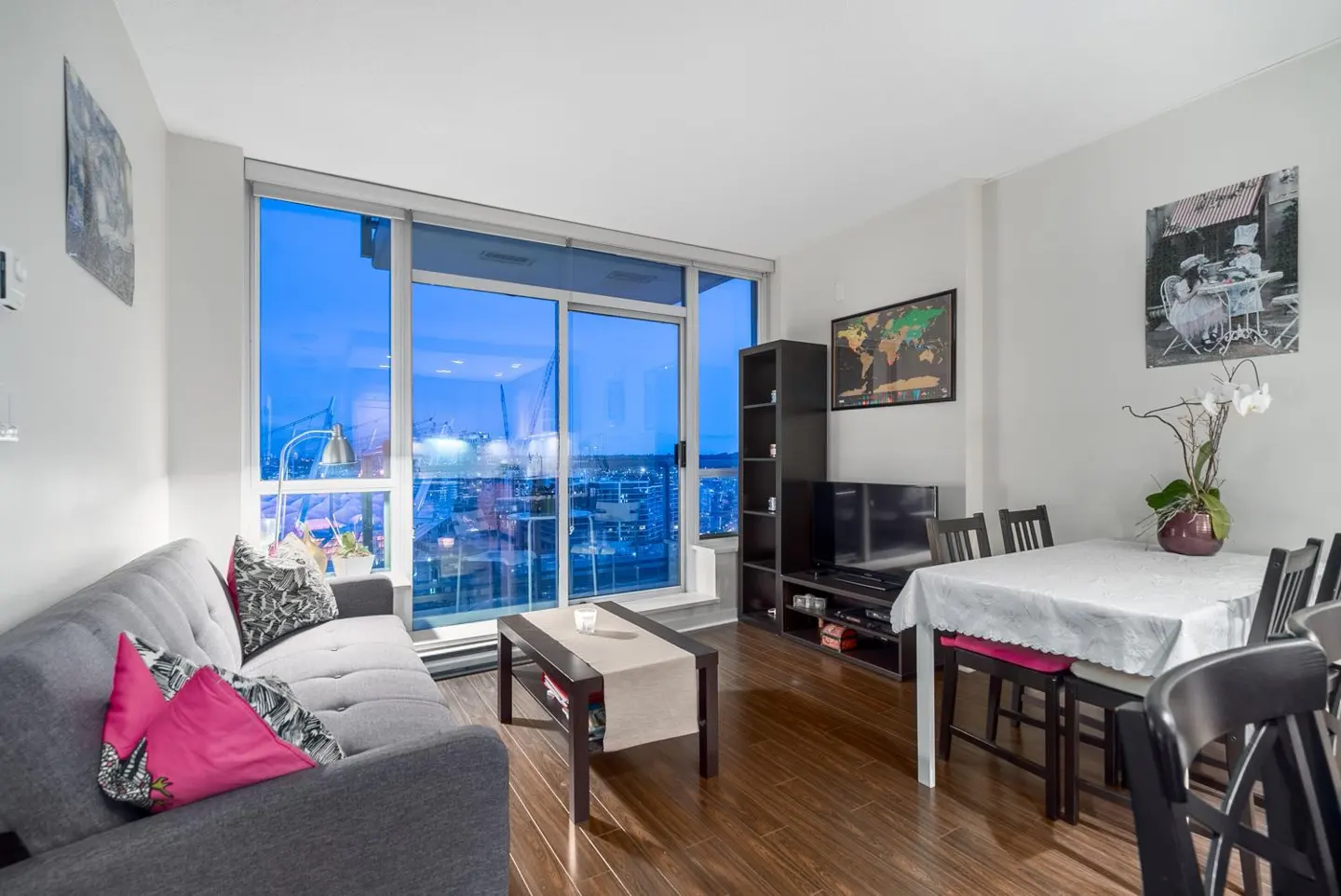 A modern apartment living room with a gray sofa, dark wood floors, and a large window overlooking a city skyline at dusk.