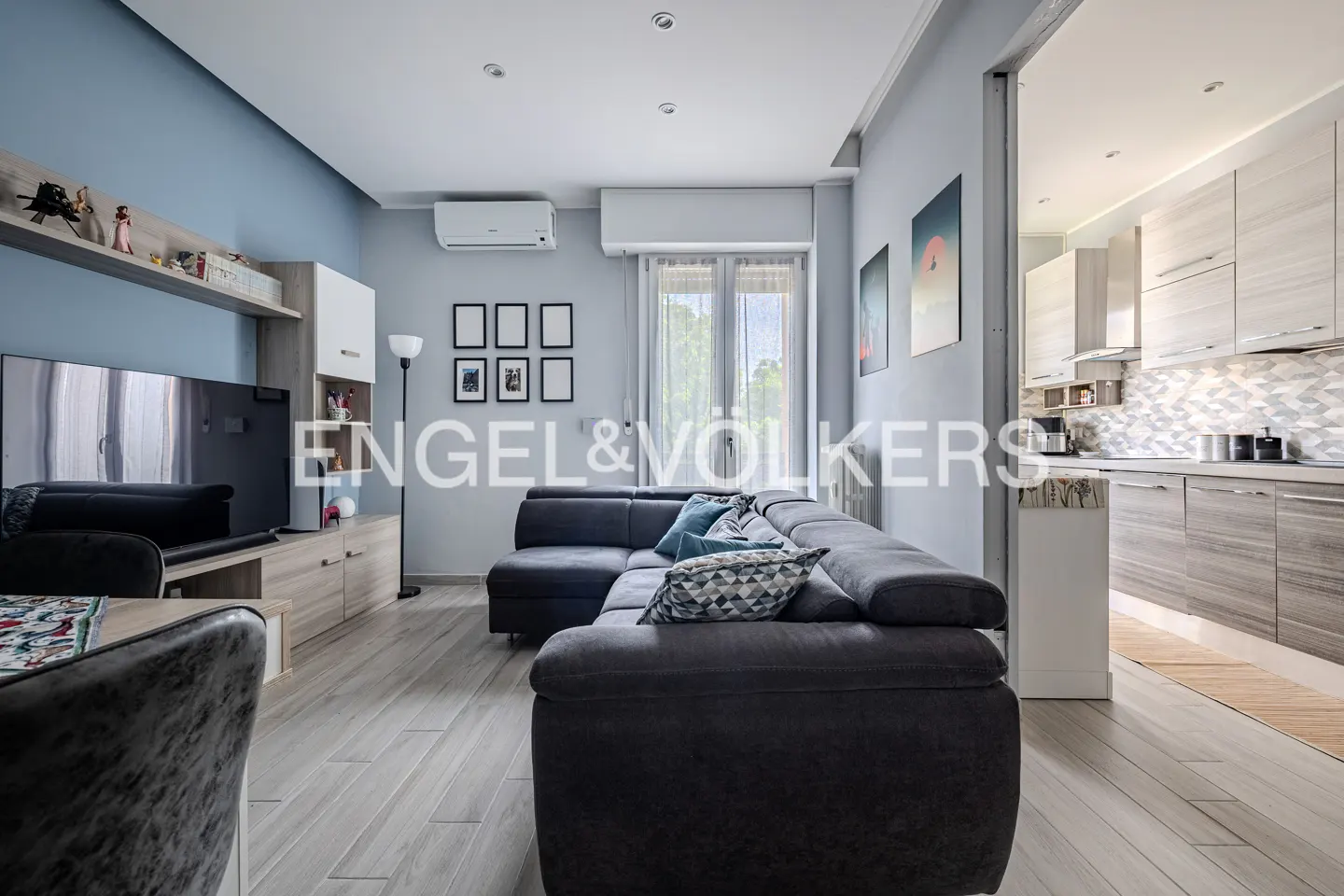 A living room with a gray sectional sofa, a TV, and a kitchen in the background. The walls are light blue.