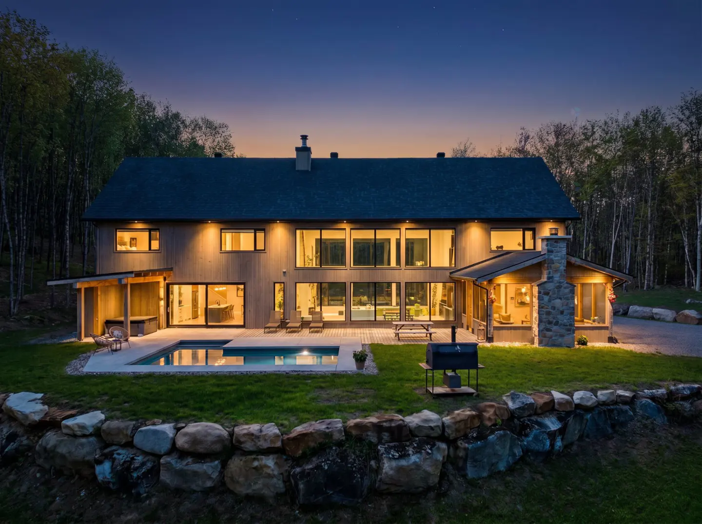 Two-story home at dusk with a pool, stone wall, and large windows. The house is surrounded by trees.