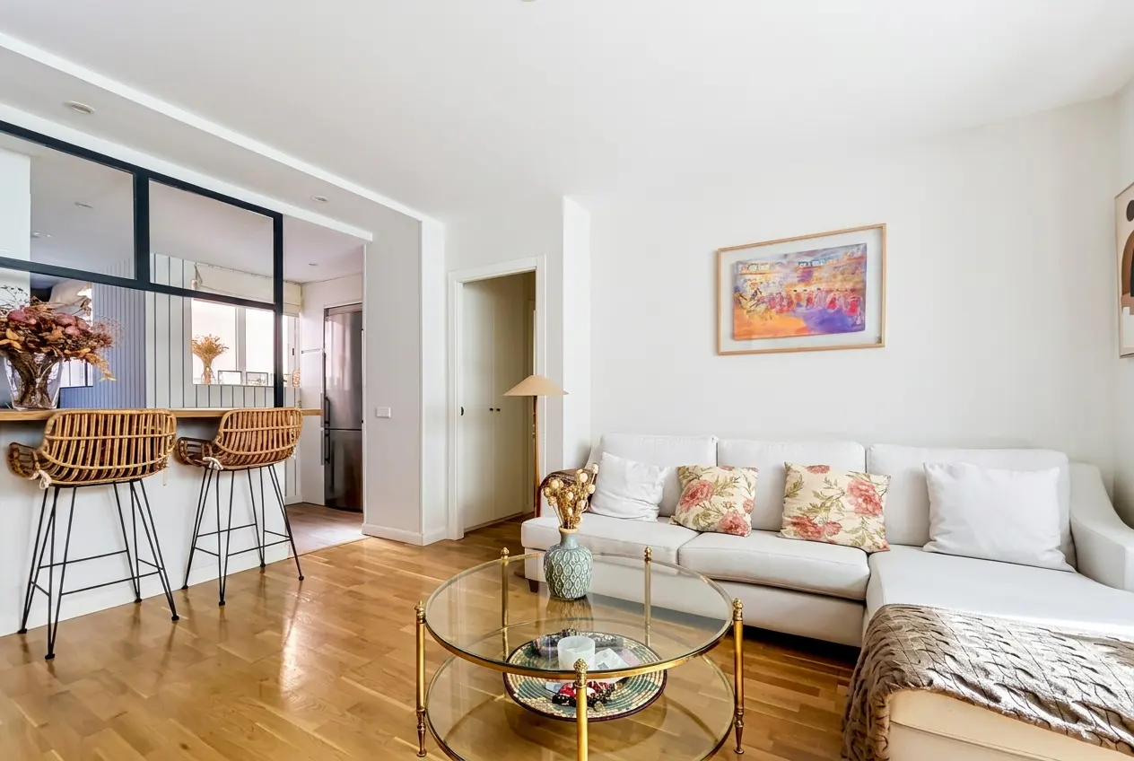 Bright living room with wood floors, white walls, and a white sectional sofa with floral pillows. A glass coffee table sits in front of the sofa. Two wicker bar stools at a counter.